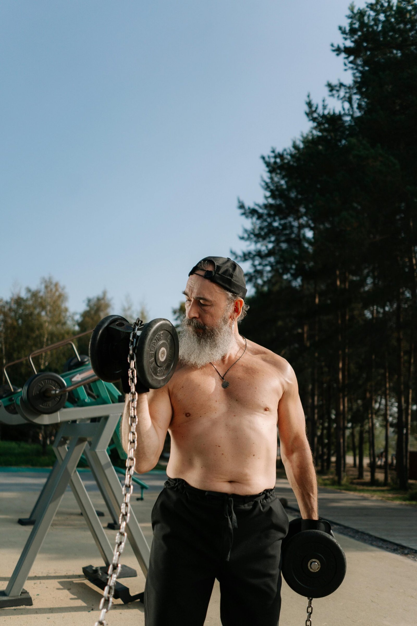 Shirtless senior man with beard lifting weights outdoors in a park.